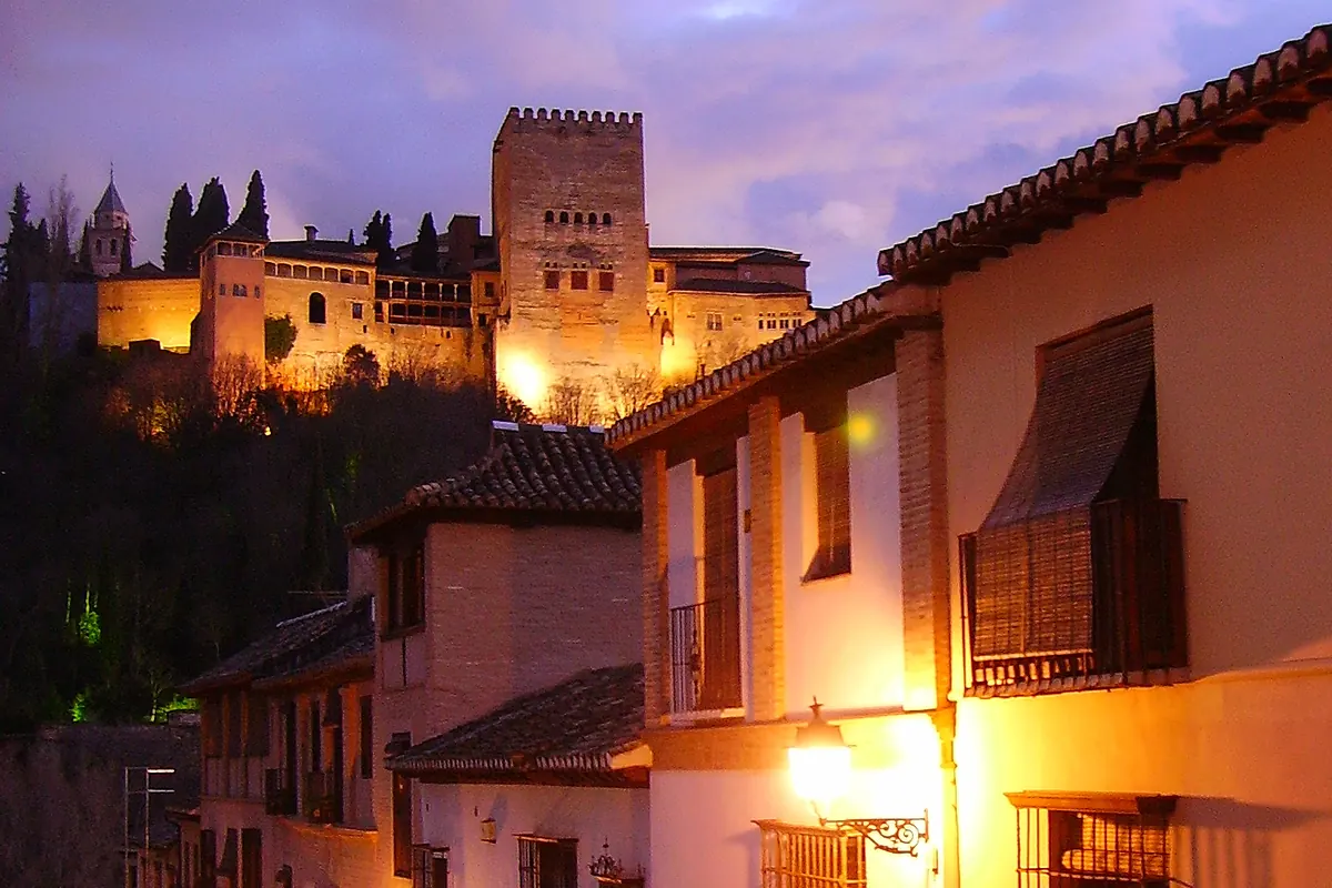 The lit towers of the Alhambra above the whitewashed houses and lamplit street of the Albaicín at dusk