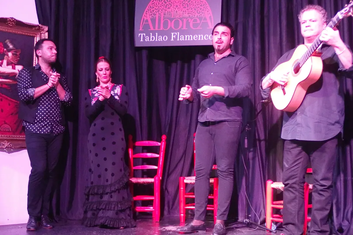 A guitarist, a singer and two clapping performers stand on the stage of La Alboreá in front of the venue sign and a framed painting of a dancer